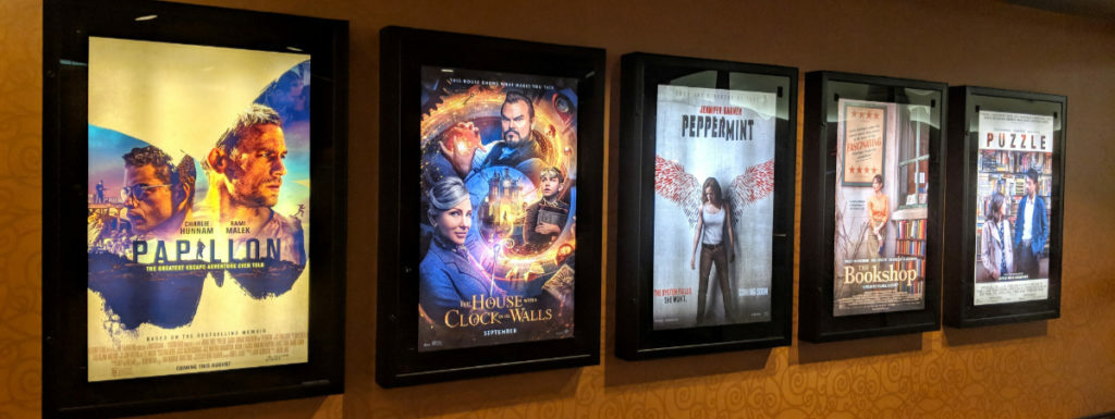 Row of movie posters including Papillon, the House with a Clock in it's Wall, Peppermint, The Bookshop, and Puzzle on a movie theatre wall